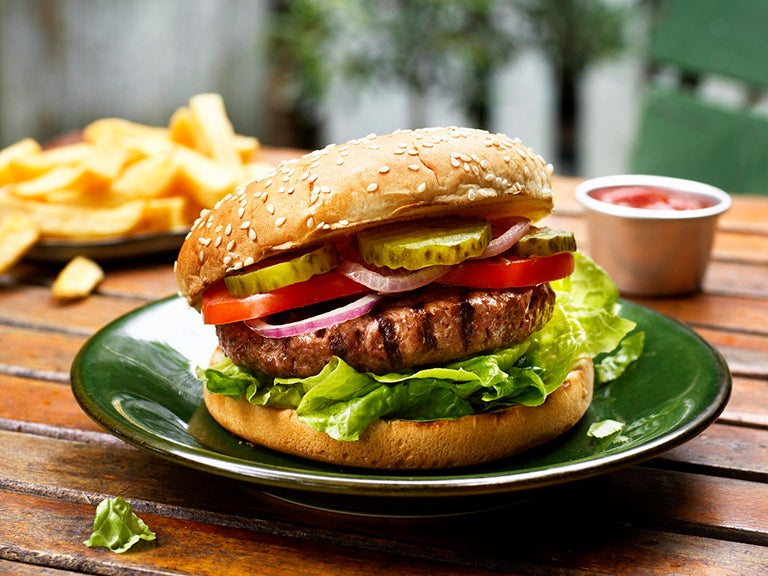 En hamburgare på en tallrik med sallad, pickles, tomat och lök. En tallrik med pommes frites i bakgrunden
