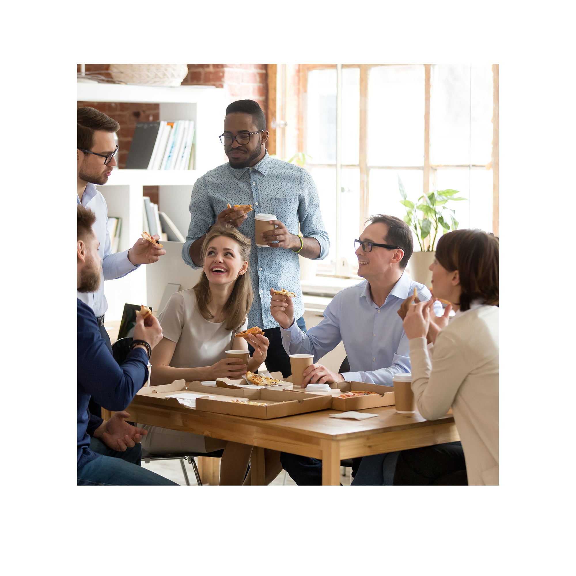 Et træbord med pizzaæsker omkring sig og omgivet af kolleger, nogle sidder, andre står og spiser pizza.