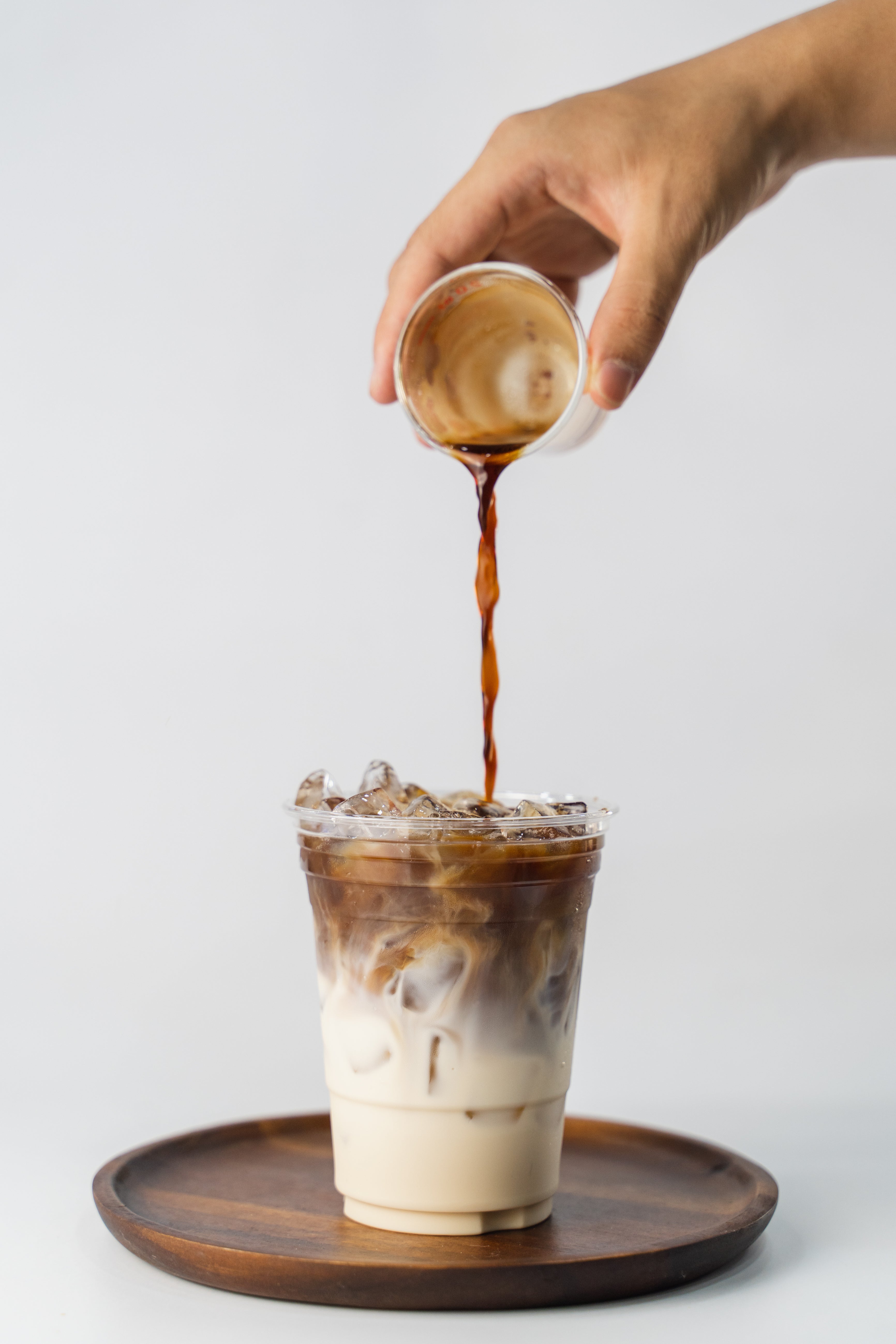 En plastmugg på en träbas med en vit bakgrund där mjölk, is och kaffe hälls i