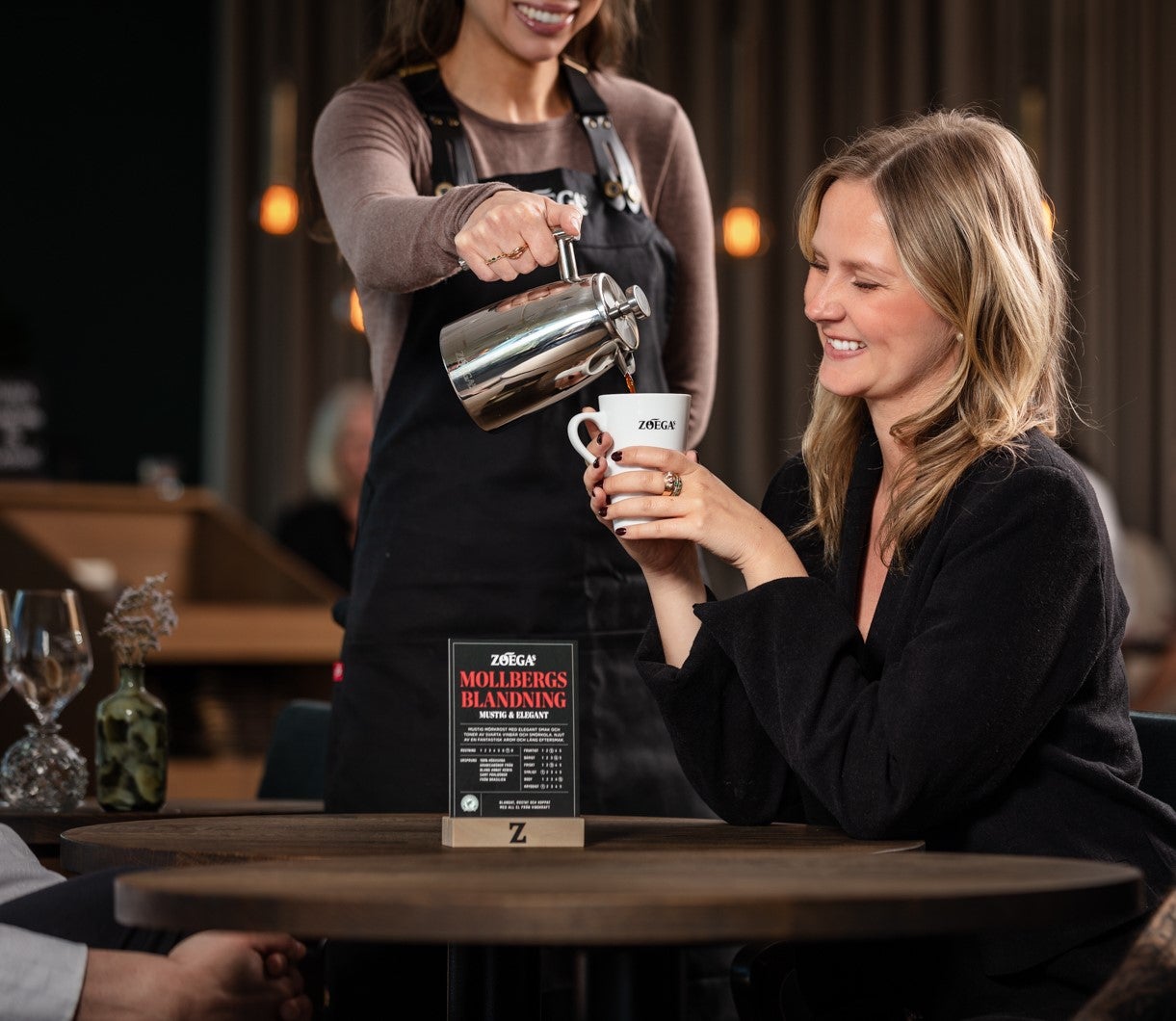 Kvinna som ler och sitter vid ett kafébord med en anställd som serverar kaffe i koppen