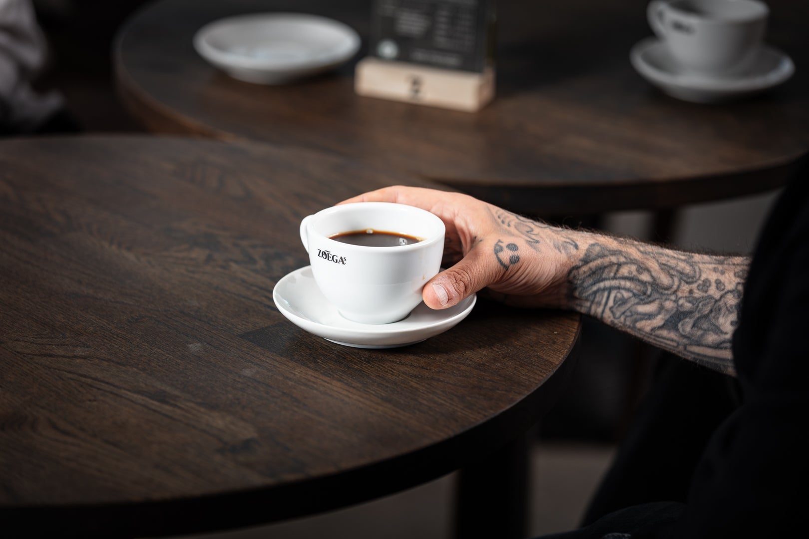 Person med tatuerad arm som tar tag i en Zoégas-kopp med kaffe