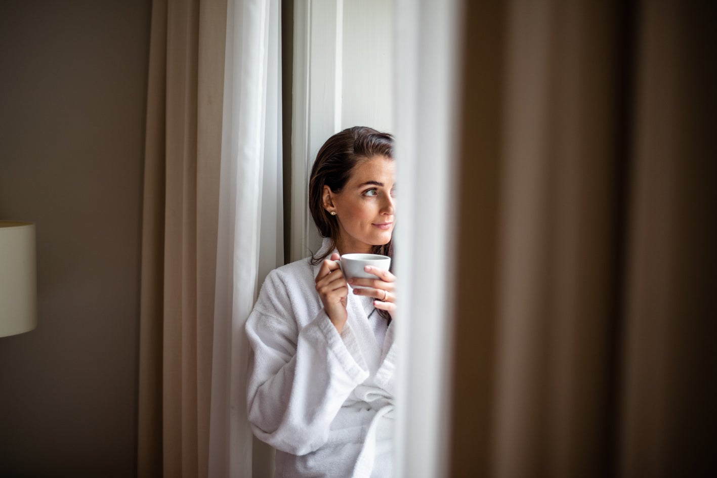 Tjej i morgonrock vid fönstret med en kopp kaffe i händerna