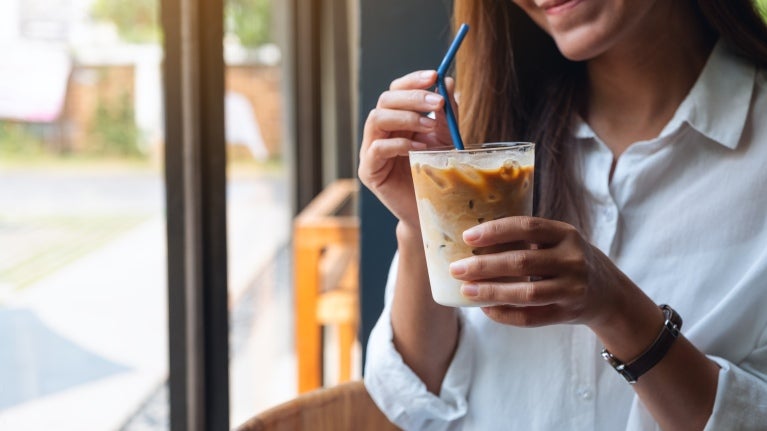 En kvinna i en vit skjorta på en kafé med en iced coffee i handen, dricker med ett sugrör