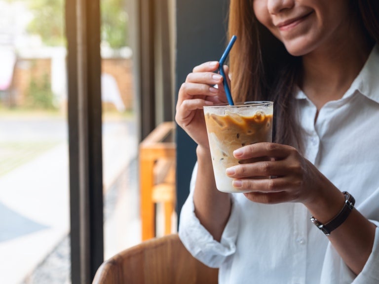 En kvinna i en vit skjorta på en kafé med en iced coffee i handen, dricker med ett sugrör