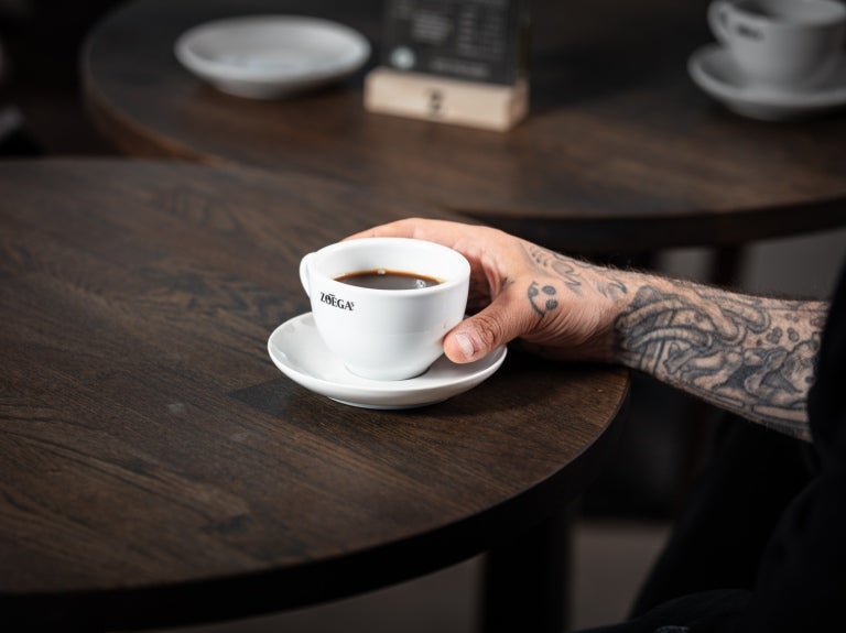Person med tatuerad arm som tar tag i en Zoégas-kopp med kaffe