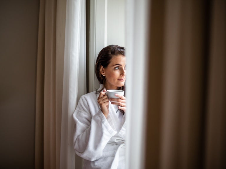 Tjej i morgonrock vid fönstret med en kopp kaffe i händerna
