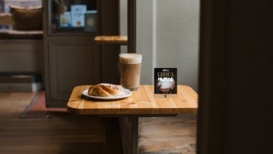 På ett bord i ett café står en latte, kanelbulle och en skylt med Zoégas varm choklad