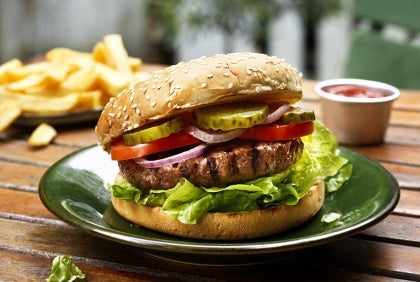 En hamburgare på en tallrik med sallad, pickles, tomat och lök. En tallrik med pommes frites i bakgrunden