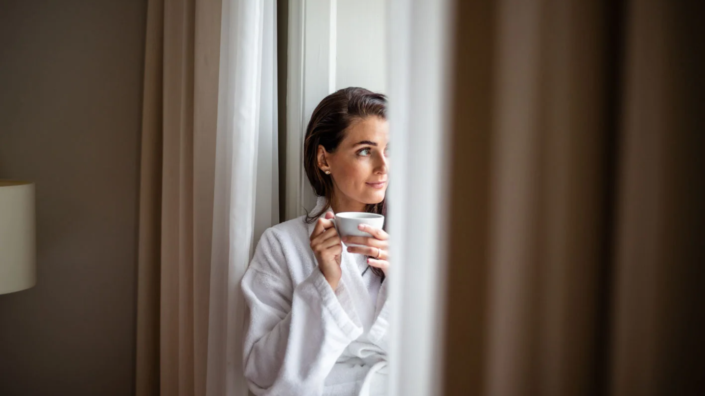 Flicka i morgonrock vid fönstret med en kopp kaffe i handen