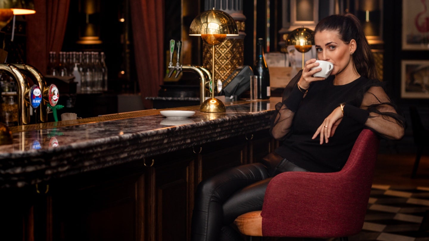 Svartklädd flicka sitter på en hög stol i en bar och njuter av sitt kaffe