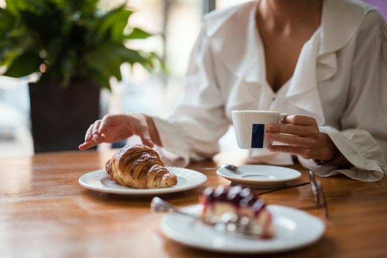Kvinna som sitter vid bordet med en kopp kaffe, croissant och en bit tårta